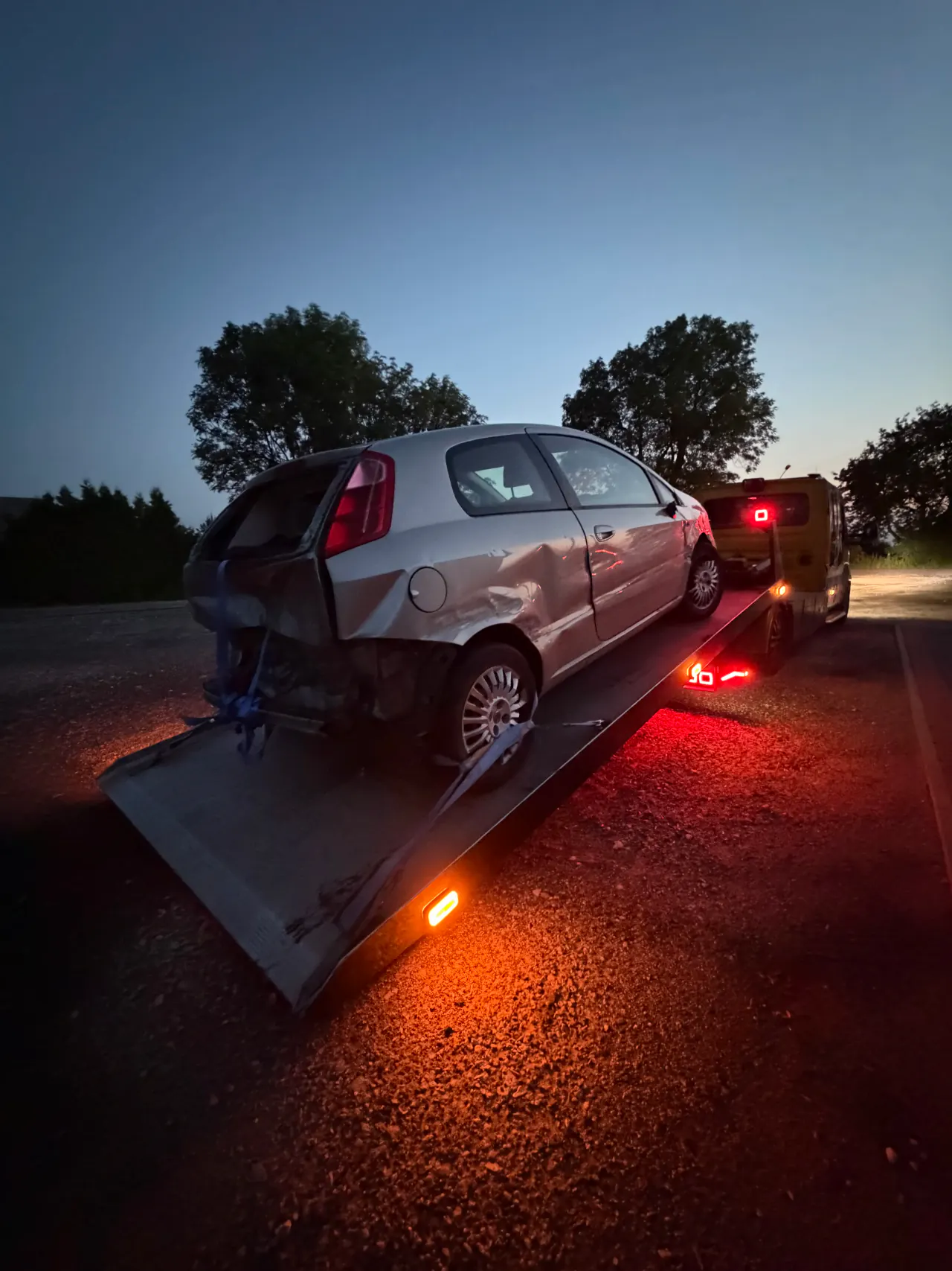 Uszkodzony srebrny samochód na opuszczonej platformie autolawety po zmroku